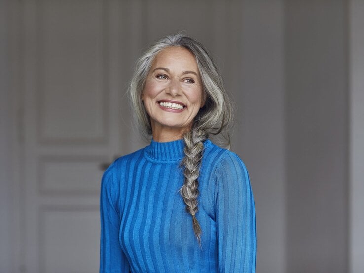 Eine lächelnde ältere Frau mit langen grauen Haaren im Zopf trägt ein blaues Kleid und steht vor einem neutralen, lichtdurchfluteten Hintergrund.