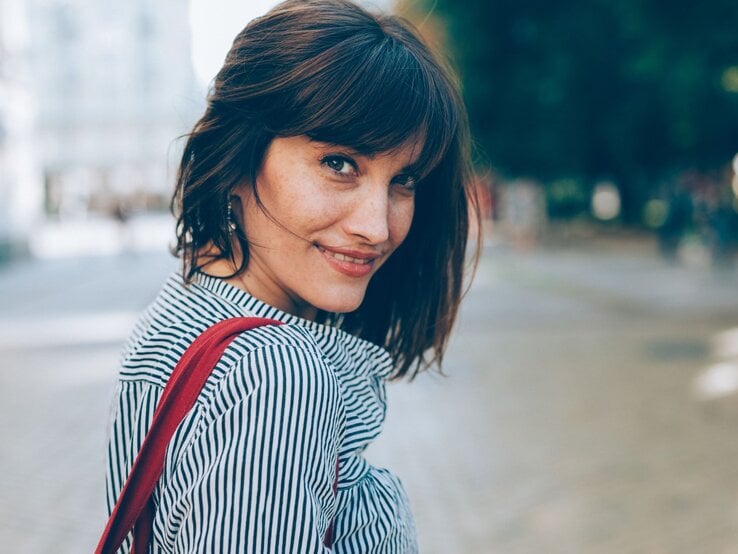 Eine brünette Frau mit Long Bob und gestreiftem Hemd blickt lächelnd über die Schulter, eine rote Tasche über der Schulter.