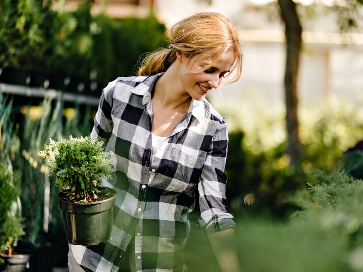 Eine blonde Frau mit lockerer Zopf-Frisur für die Gartenarbeit trägt eine Pflanze in einem Blumentopf, umgeben von üppigem Grün.