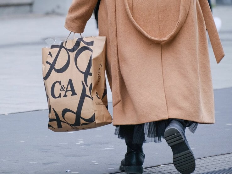 Eine Person geht auf einer Straße und hält eine C&A-Einkaufstüte. Sie trägt einen stilvollen, kamelfarbenen Mantel und schwarze Stiefel, was auf eine kalte Jahreszeit hindeutet. Der Fokus des Fotos liegt auf der Einkaufstüte und der unteren Hälfte der Person, während der obere Teil und der Hintergrund unscharf bleiben. Dies vermittelt das Gefühl des Einkaufens in der Stadt und der Bewegung im Alltagsleben.