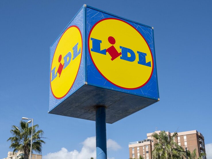 Das Foto zeigt ein großes, würfelförmiges Werbeschild von Lidl an einem klaren, sonnigen Tag. Auf jeder sichtbaren Seite des Würfels ist das gelbe Lidl-Logo mit dem roten Schriftzug auf blauem Hintergrund zu sehen. Im Hintergrund des Schildes sind Palmen und ein Gebäude, was darauf hindeutet, dass sich der Standort in einem städtischen Gebiet mit warmem Klima befindet.