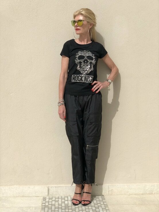  Frau, die ein schwarzes T-Shirt mit einem Totenkopf-Design und der Aufschrift "Rock N' Roll" trägt. Sie kombiniert dies mit schwarzen Nylon-Fallschirmhosen und schwarzen High-Heels. | © Renate Zott