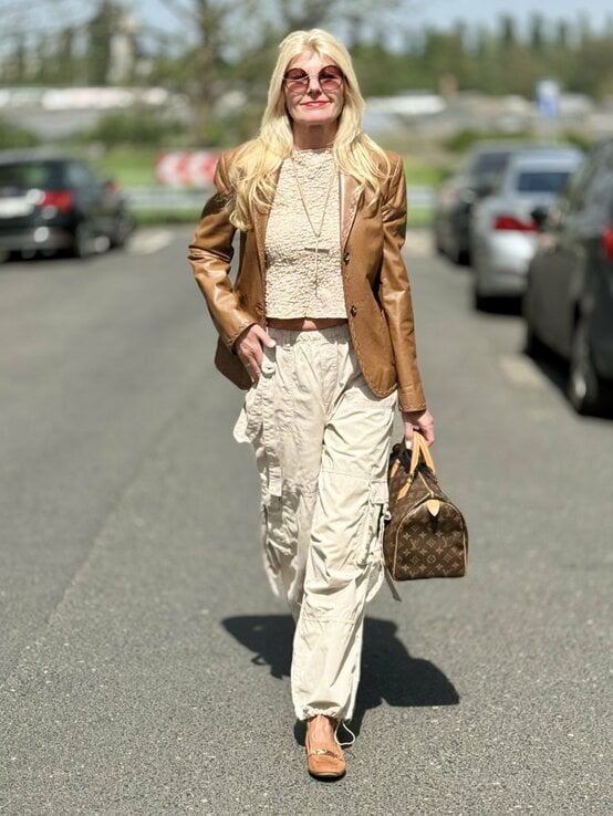 Frau, die einen eleganten, braunen Lederblazer trägt, kombiniert mit einem beigen, strukturierten Oberteil und cremefarbenen Fallschirmhosen. Sie trägt dazu flache, braune Schuhe und eine Designer-Handtasche. | © Renate Zott