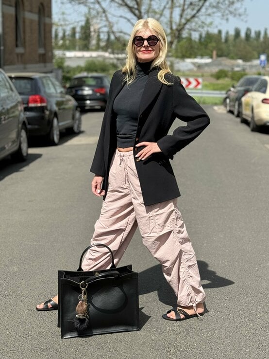 Frau, die einen schwarzen Blazer und ein schwarzes Oberteil trägt, kombiniert mit rosa Fallschirmhosen. Sie trägt eine große, schwarze Tasche und schwarze Sandalen. | © Renate Zott 