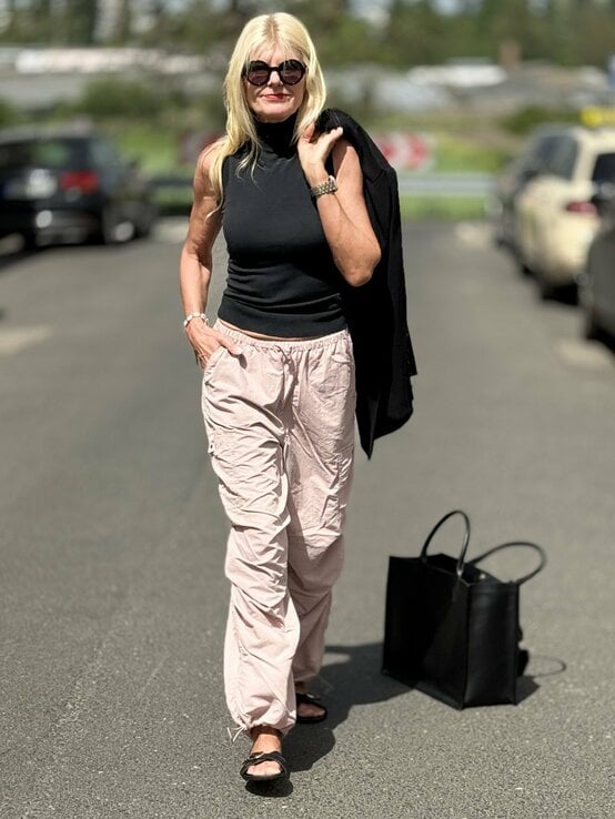  Frau, die ein ärmelloses, schwarzes Oberteil trägt, kombiniert mit rosa Fallschirmhosen. Sie trägt eine Sonnenbrille und hält einen schwarzen Blazer lässig über der Schulter. | © Renate Zott