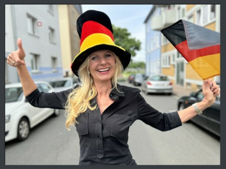 Frau, die eine große, farbenfrohe Hut in den Farben der deutschen Flagge trägt und eine Deutschlandflagge in der Hand hält. Sie lächelt und zeigt einen Daumen hoch, während sie auf einer Straße steht.
