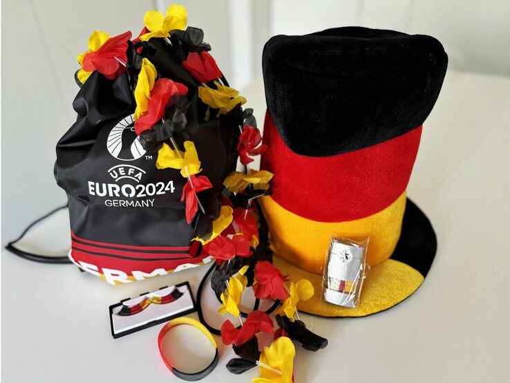 Sammlung von Fan-Accessoires für die EM 2024 in Deutschland. Zu den gezeigten Artikeln gehören ein großer Hut in den Farben der deutschen Flagge, eine Blumenkette in Schwarz-Rot-Gold, ein Armband in den gleichen Farben, und ein Rucksack mit dem Logo der UEFA Euro 2024. | © Renate Zott