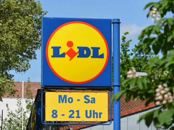 Werbeschild von Lidl mit gelbem Logo und Öffnungszeiten, im Hintergrund grünes Laub und blauer Himmel.