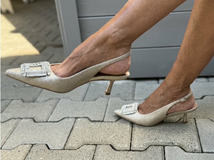 Beine, die in glitzernden Slingback-Pumps stecken. Diese Schuhe haben spitze Kappen und sind mit funkelnden Verzierungen, wahrscheinlich Strasssteinen, auf der Vorderseite dekoriert. Die Schuhe haben einen schmalen, mittelhohen Absatz und einen Fersenriemen, der um die Ferse verläuft.  | © Renate Zott