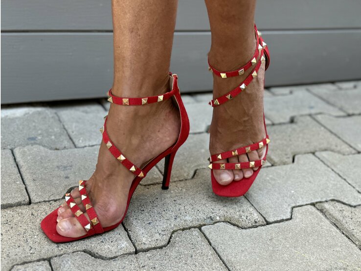 Beine, die in roten High Heels mit goldenen Nieten stecken. Diese Schuhe haben mehrere dünne Riemen, die den Fuß und den Knöchel umschließen, und einen hohen Stilettoabsatz. Die goldenen Nieten sind gleichmäßig auf den Riemen verteilt und verleihen den Schuhen einen auffälligen und stilvollen Look. | © Renate Zott