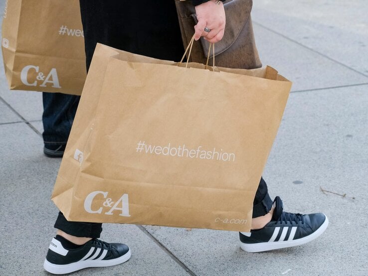 Zwei Personen gehen mit großen braunen C&A-Einkaufstaschen und tragen sportliche Schuhe auf einem grauen Gehweg.
