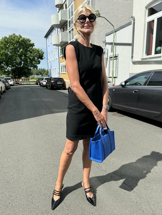 Frau steht auf einer Straße und trägt ein elegantes schwarzes Kleid. Sie hat eine blaue Handtasche in der Hand und trägt schwarze, spitze Schuhe mit Riemchen. Die Frau trägt eine Sonnenbrille und ihre blonden Haare sind locker hochgesteckt.  | © Renate Zott