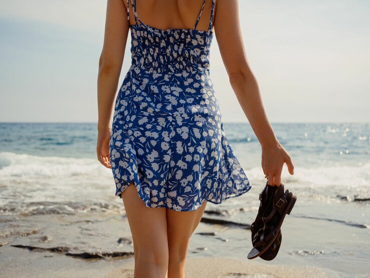Frau in geblümtem Kleid spaziert barfuß mit Sandalen in der Hand an der Küste entlang bei ruhigem Meer.