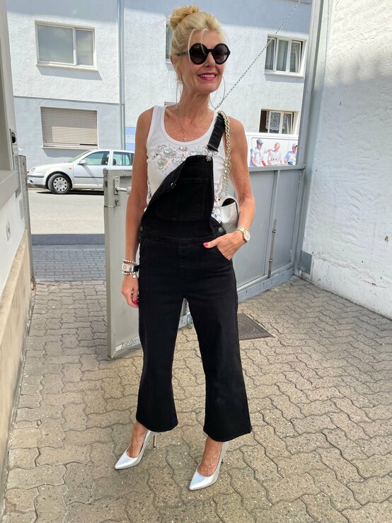 Frau, die lächelnd in einer städtischen Umgebung steht. Sie trägt eine schwarze Latzhose über einem weißen, verzierten Tanktop und silberne High Heels. Dazu kombiniert sie eine große Sonnenbrille und trägt eine kleine Handtasche mit einer Kette als Trageriemen. Ihr Haar ist zu einem lockeren Dutt hochgesteckt. | © Renate Zott