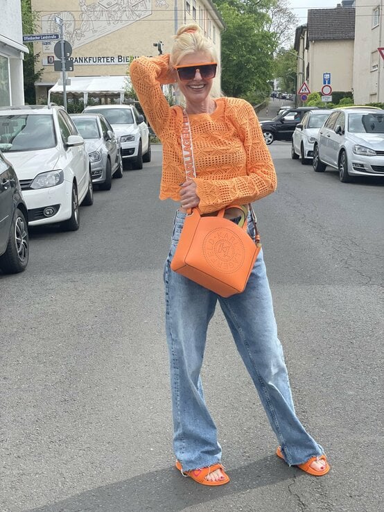 Frau, die fröhlich auf einer Straße steht. Sie trägt ein leuchtend oranges, gestricktes Oberteil, eine große orangefarbene Sonnenbrille, eine orange Tasche mit einem breiten Riemen, weite blaue Jeans und orange Sandalen. Ihr Haar ist hochgebunden. | © Renate Zott