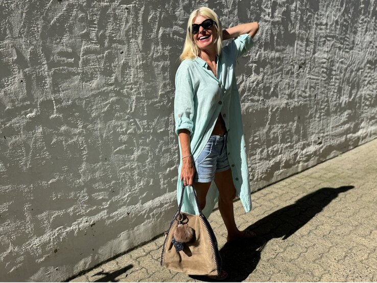 Frau mit blonden Haaren und Sonnenbrille, die sich leger an einer grauen Wand im Freien anlehnt. Sie trägt ein hellblaues, offenes Hemd über einem weißen Top und blaue Denim-Shorts. Ihr Outfit wird durch eine große beige Handtasche mit Fransen abgerundet.