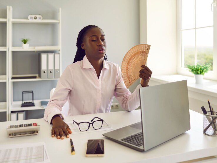 Geschäftsfrau im weißen Hemd kühlt sich im Büro mit einem Fächer, während sie am Laptop arbeitet.