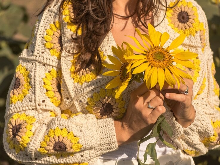 Nahaufnahme einer Frau in einem gestrickten Sonnenblumenpullover, die zwei große gelbe Sonnenblumen in den Händen hält.