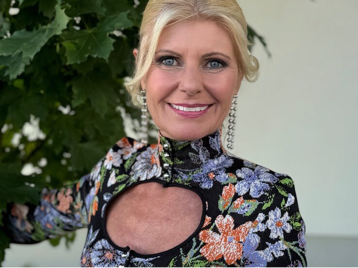 Ältere Frau mit einem strahlenden Lächeln, die in die Kamera blickt. Sie hat blondes Haar und trägt ein farbenfrohes Kleid mit einem floralen Muster und einem auffälligen Ausschnitt am Dekolleté.