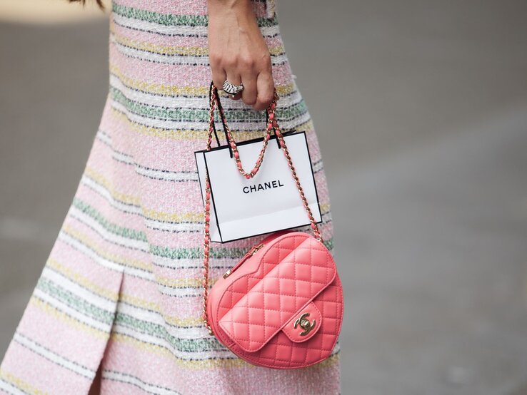 Frau in pastellfarbenem gestreiftem Rock hält eine rosa Chanel-Tasche und eine Chanel-Papiertüte.