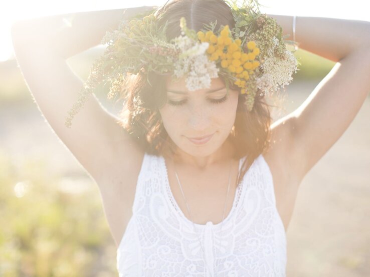 Eine Frau mit Blumenkranz und weißem Spitzenkleid steht verträumt im Sonnenlicht mit erhobenen Armen.