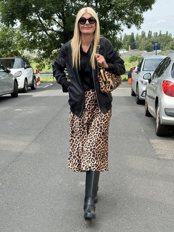 Frau mittleren Alters läuft auf einem Parkplatz und trägt einen eleganten Look: eine schwarze Bomberjacke über einer schwarzen Bluse, kombiniert mit einem leopardenmuster Rock und schwarzen Stiefeln. | © Renate Zott