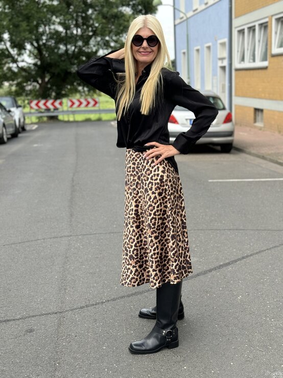 Frau mittleren Alters von der Seite auf einem Parkplatz. Sie trägt eine schwarze Bomberjacke über einer schwarzen Bluse, kombiniert mit einem leopardenmuster Rock und schwarzen Stiefeln. | © Renate Zott