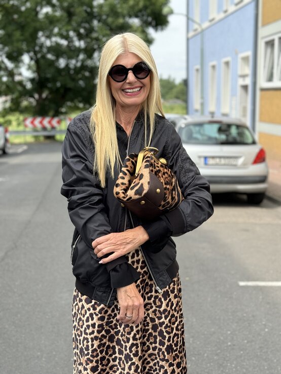 Stilvolle Frau mittleren Alters posiert auf einer Straße, lächelnd in die Kamera. Sie trägt eine schwarze Bomberjacke, darunter eine schwarze Bluse und einen leopardenmuster Rock.  | © Renate Zott