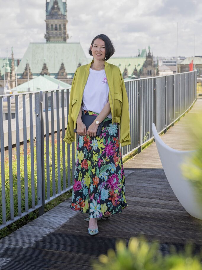 Frau mit kurzen schwarzen Haaren, gelber Jacke, weißem T-Shirt und buntem Blumenrock steht lächelnd auf einer Dachterrasse vor dem Hamburger Rathaus. | © Bild der Frau/ Ulrike Schacht