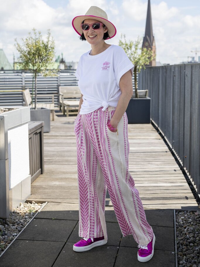 Frau mit Strohhut, rosa Sonnenbrille, weißem T-Shirt und rosa-weiß gemusterten Hosen steht lächelnd auf einer Dachterrasse. | © Bild der Frau/ Ulrike Schacht