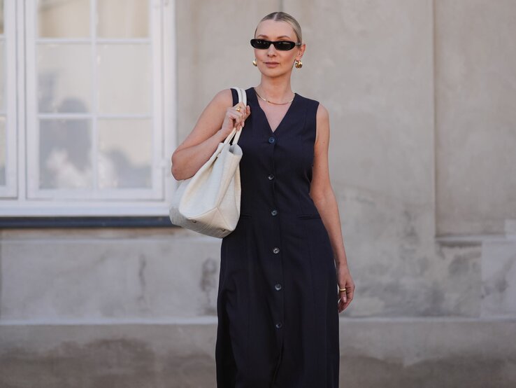 Frau in schwarzem Kleid und Sonnenbrille trägt weiße Tasche, steht vor einem hellen Fenster und einer beige Wand.