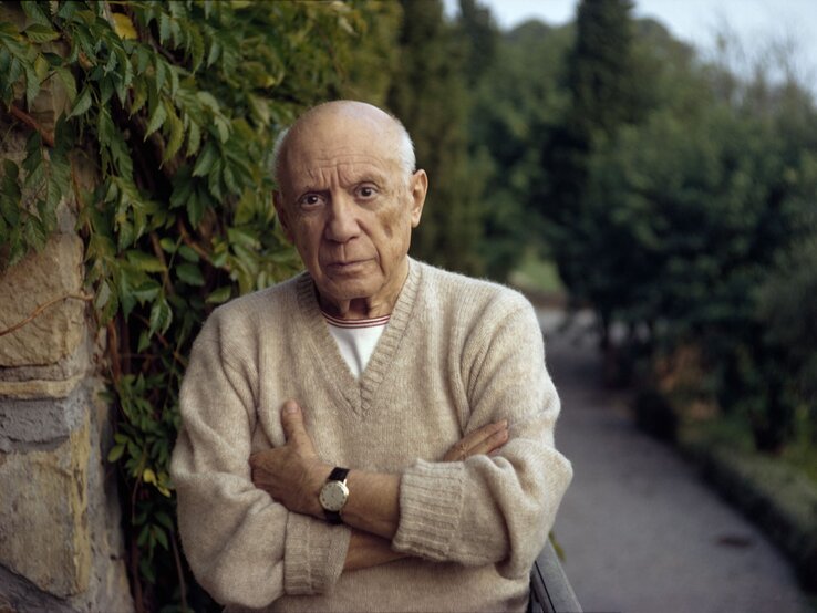 Pablo Picasso trägt einen beigen Pullover und steht mit ernster Miene vor einer Mauer mit grünen Pflanzen.