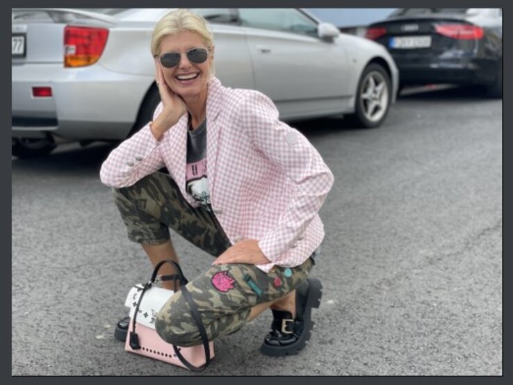 Lächelnde Frau mittleren Alters mit blonden Haaren und Sonnenbrille, die in einer Hockstellung auf einem Parkplatz posiert. Sie trägt eine rosafarbene, karierte Jacke, ein schwarzes T-Shirt mit Aufdruck, Tarnhosen und schwarze Schuhe.