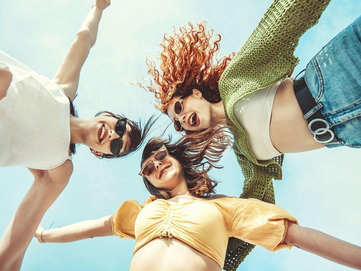 Lächelnde Frauen in sommerlicher Kleidung und Sonnenbrillen, von unten fotografiert, mit ausgestreckten Armen unter blauem Himmel.