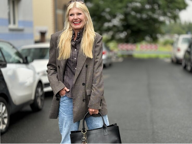 Frau mit langen blonden Haaren, die einen karierten Blazer und eine gepunktete Bluse trägt. Sie kombiniert das Outfit mit einer hellblauen Jeans.