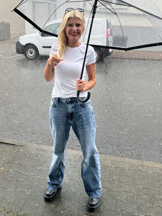 Frau mit langen blonden Haaren, die lächelnd in die Kamera blickt. Sie trägt ein schlichtes weißes T-Shirt und eine hellblaue Jeans. In ihrer Hand hält sie einen Regenschirm. | © Renate Zott