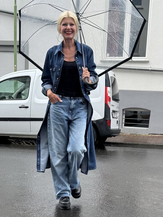 Frau mit blonden Haaren, die einen durchsichtigen Regenschirm hält. Sie trägt einen langen, offenen Jeansmantel über einem schwarzen Oberteil und einer weiten hellen Jeans. | © Renate Zott