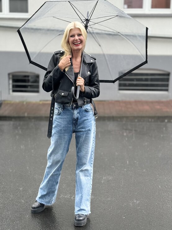 Frau mit blonden Haaren, die einen durchsichtigen Regenschirm hält. Sie trägt eine schwarze Lederjacke über einem dunklen Oberteil und kombiniert dies mit einer hellen, weit geschnittenen Jeans. | © Renate Zott