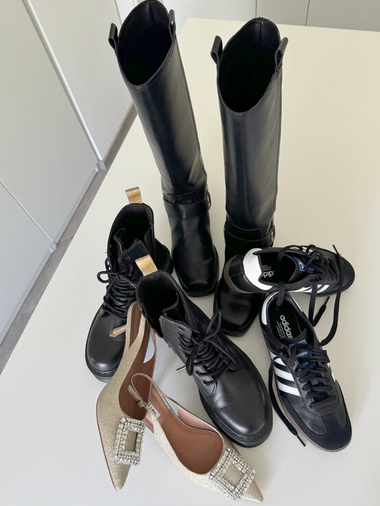 Auswahl verschiedener Schuhe, die auf einem weißen Tisch platziert sind. Es sind schwarze kniehohe Stiefel, schwarze Schnürstiefeletten, schwarze Sneaker von Adidas mit weißen Streifen sowie ein Paar elegante, goldene Slingback-Pumps | © Renate Zott