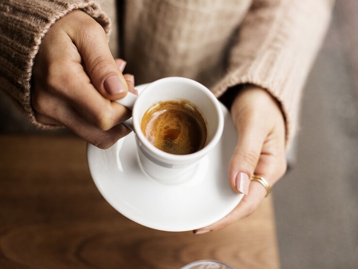 Nahaufnahme von zwei Händen mit gepflegten Nägeln, die eine weiße Espressotasse auf einem Holztisch halten.