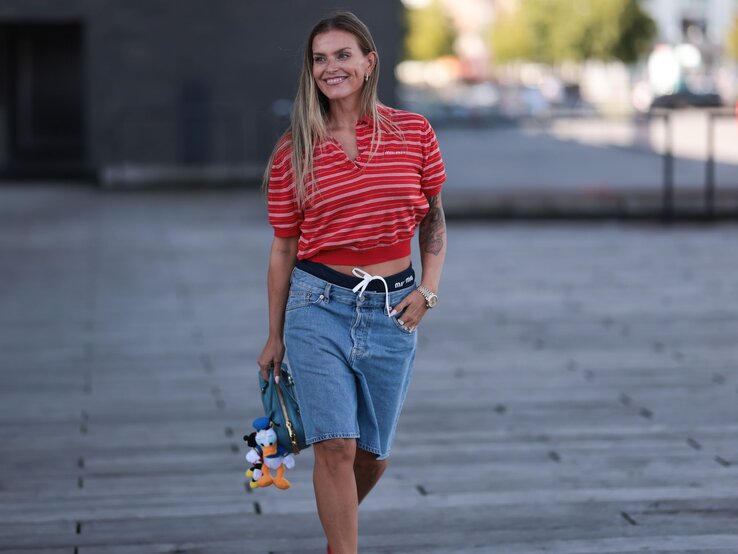 Lächelnde Frau in sommerlichem Outfit mit Jeansshorts, rot gestreiftem Shirt und Tätowierung am rechten Arm, hält eine blaue Tasche mit Stoffanhängern.