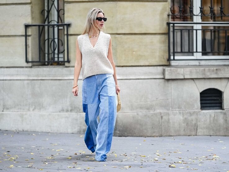 Junge blonde Frau in beigefarbenem Stricktop und weiter Patchwork-Jeans geht mit kleiner Tasche auf herbstlicher Straße.