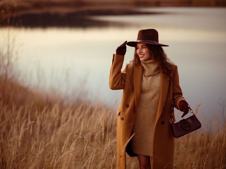 Zu sehen ist eine brunette Frau. Sie trägt einen Hut, Handschuhe, Mantel und Strickkleid in herbstlicher Atmossphäre.