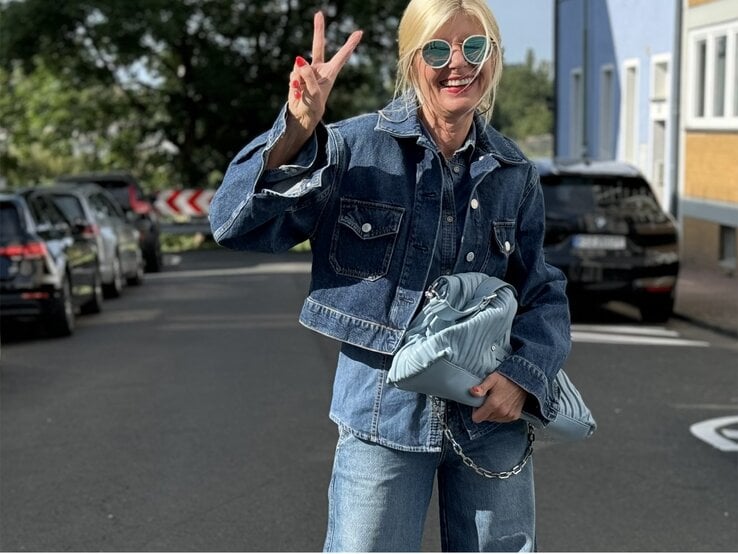 Blonde Frau in einem modischen Jeans-auf-Jeans-Outfit. Sie trägt eine Sonnenbrille und zeigt ein fröhliches Lächeln, während sie ein Peace-Zeichen mit der Hand macht.