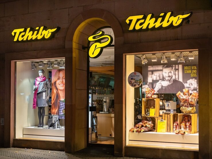 Außenansicht einer Tchibo-Filiale mit gelbem Logo, beleuchtetem Schaufenster, das Kleidung und Kaffeeprodukte präsentiert.