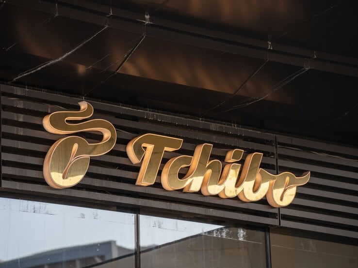 Goldenes Tchibo-Logo in geschwungener Schrift auf dunklem Ladenhintergrund mit reflektierender Glasfassade.