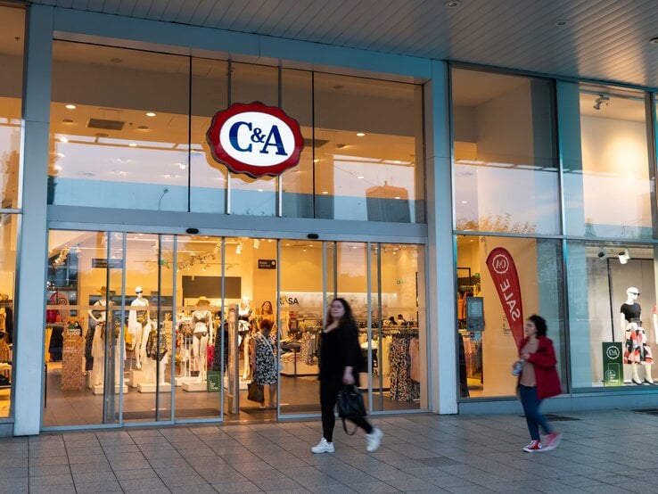 C&A-Geschäft mit großen Glasfenstern, leuchtendem Logo, Schaufensterpuppen und vorbeigehenden Passanten bei Abenddämmerung.