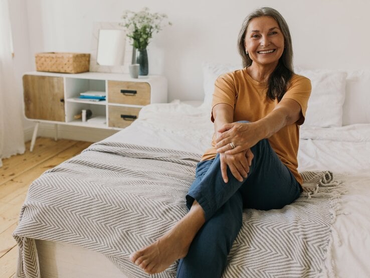 Ältere Frau mit grauen langen Haaren sitzt lächelnd mit Straßenkleidung auf einem Bett in einem hellen Zimmer mit schlichter Dekoration.