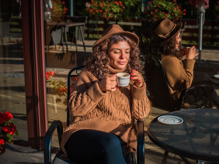 Zu sehen ist eine Frau mittleren Alters, die einen braunen Hut und einen braunen Zopfstrickpullover trägt, während sie Kaffee trinkt