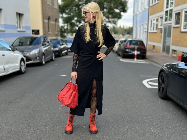 Frau in einem auffälligen Outfit auf der Straße. Sie trägt ein schwarzes Kleid mit Spitzenärmeln und einem hohen Beinschlitz. Dazu kombiniert sie rote Stiefel und eine rote Handtasche, die dem Look farbliche Akzente verleihen. Die Frau trägt ihr blondes Haar offen und eine Sonnenbrille.